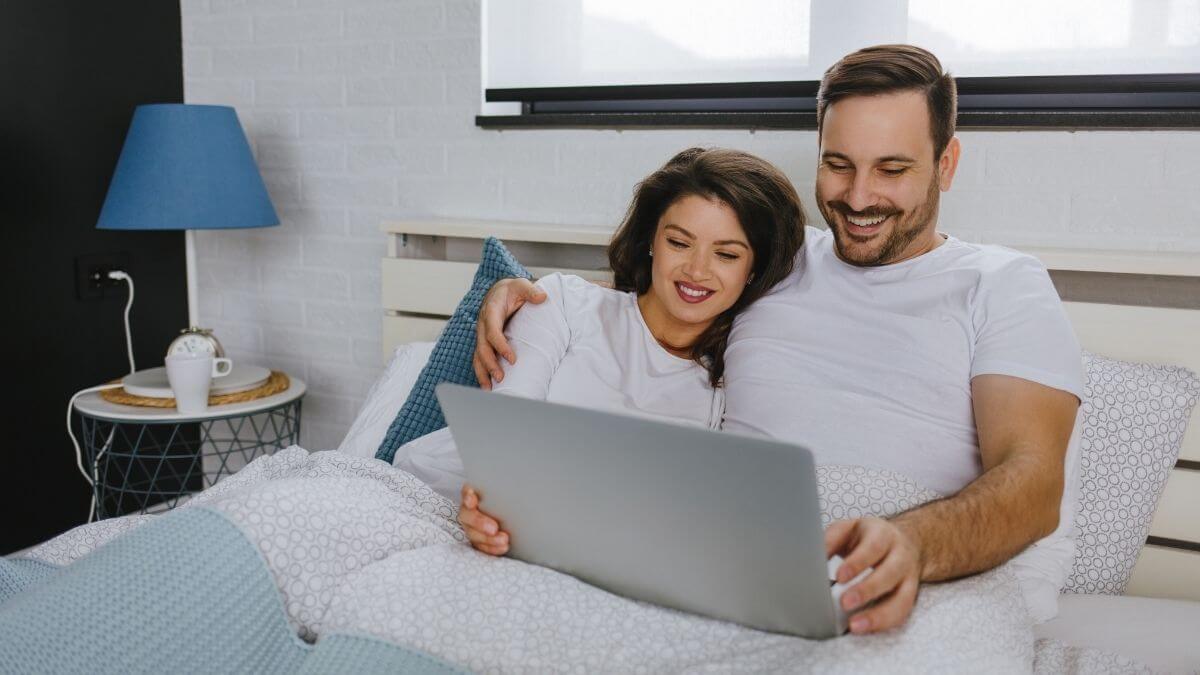 A couple smiling and looking at a laptop together while relaxing in bed