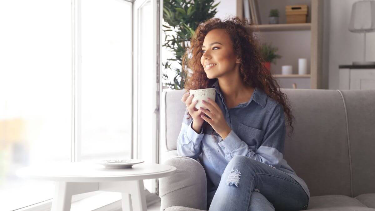 A woman sitting in an armchair holding a coffee cup and gazing out the window with a smile